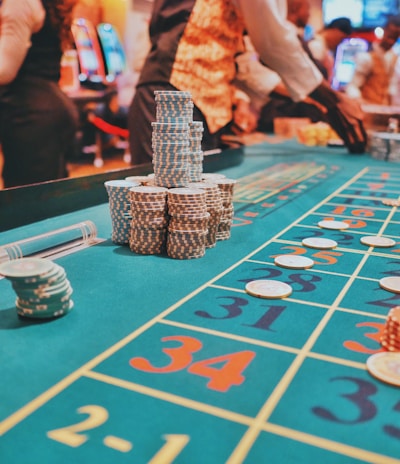 A casino scene with a roulette table features prominently, covered in green felt with colorful numbers and betting squares. Several stacks of poker chips are placed on different numbers, and the hands of several people are visible as they interact with the table. In the background, there are slot machines and more people, creating a vibrant and busy atmosphere.