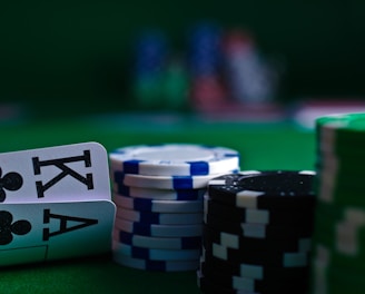 A casino scene featuring two playing cards, the ace and king of clubs, partially concealed by a player's hand. Nearby, there are stacks of multicolored poker chips on a green felt table, with blurred colorful background elements suggesting other chip stacks or cards.