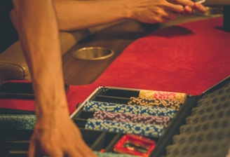A person reaches into a poker chip case on a red felt table, while another person holds playing cards. The scene is dimly lit, creating a warm and engaged atmosphere.