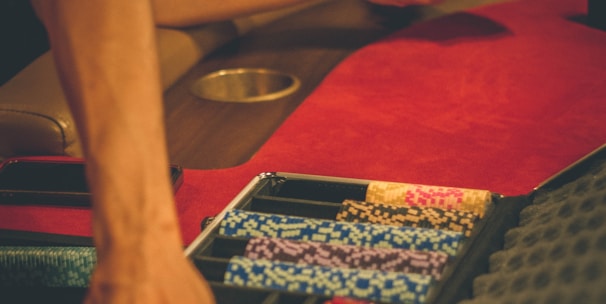 A person reaches into a poker chip case on a red felt table, while another person holds playing cards. The scene is dimly lit, creating a warm and engaged atmosphere.