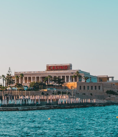 A seaside view of a large building labeled as a casino, situated on a rocky shore. The building is surrounded by palm trees and other smaller structures. The calm sea water reflects the sky, and there are several closed umbrellas lined along the waterfront.