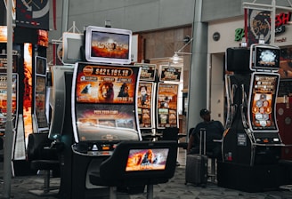 A collection of slot machines is situated inside a building with bright, colorful screens displaying various games and gaming themes. A person is seated nearby, seemingly relaxed with luggage beside them, and the atmosphere is lively with electronic displays.