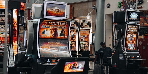 A collection of slot machines is situated inside a building with bright, colorful screens displaying various games and gaming themes. A person is seated nearby, seemingly relaxed with luggage beside them, and the atmosphere is lively with electronic displays.